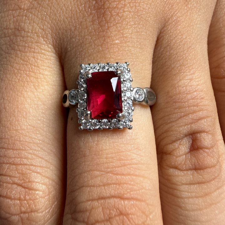A close-up of a ring worn on a finger, featuring a rectangular red gemstone surrounded by a halo of small clear stones on a silver-toned band.
