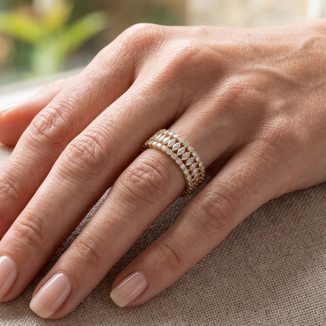 A close-up of a hand wearing a delicate gold ring with rows of small clear stones, resting on a textured beige fabric surface.