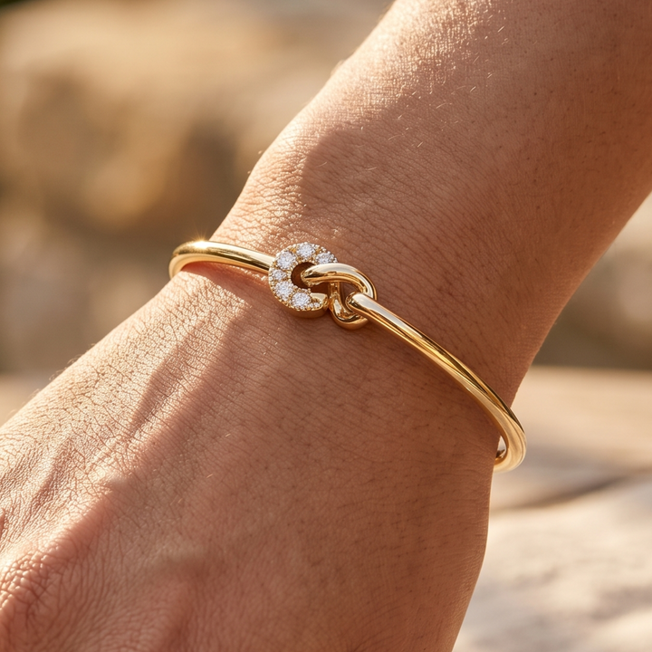 A close-up of a gold bangle bracelet with a knot motif set with small clear stones, worn on a person's wrist against a warm, blurred outdoor background.