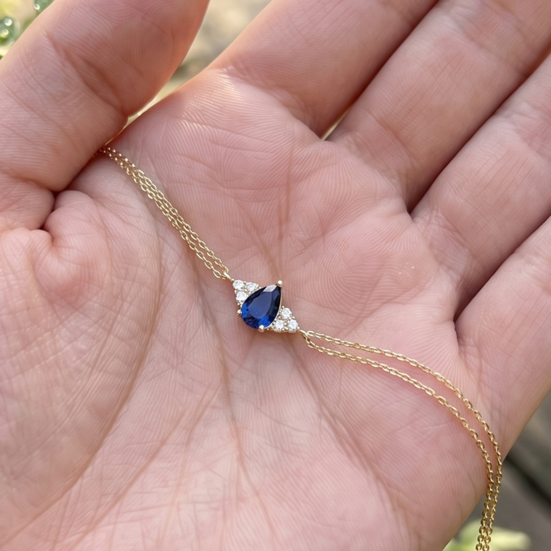 A delicate gold chain necklace with a pear-shaped blue gemstone pendant, held in a person's open palm.