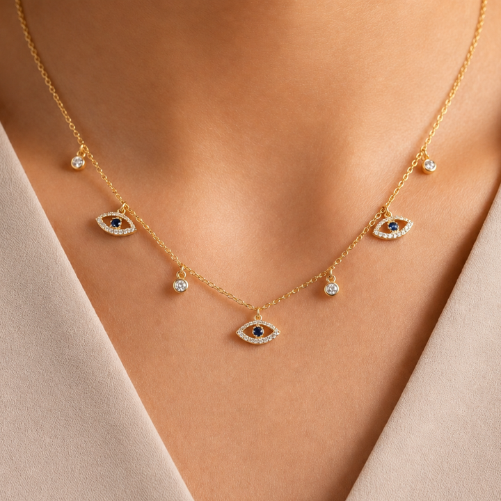 A gold necklace with multiple evil eye charms and small clear stone accents, shown worn on a model's neck against a soft beige top.