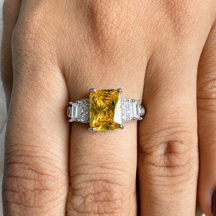 A yellow cushion-cut emerald diamond ring with baguette side stones in a silver setting, shown worn on a person's finger.