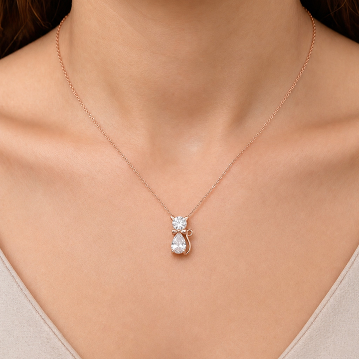 A close-up of a person's neck and chest wearing a delicate rose gold necklace with a pendant shaped like a small cat holding a pear-shaped clear gemstone.