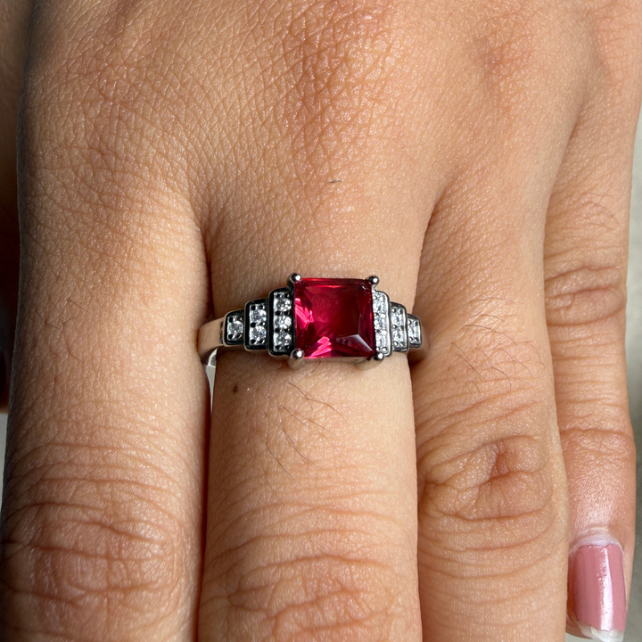 A close-up of a ring worn on a finger, featuring a large square-cut pinkish-red center stone with small clear stones on either side of a silver-toned band.