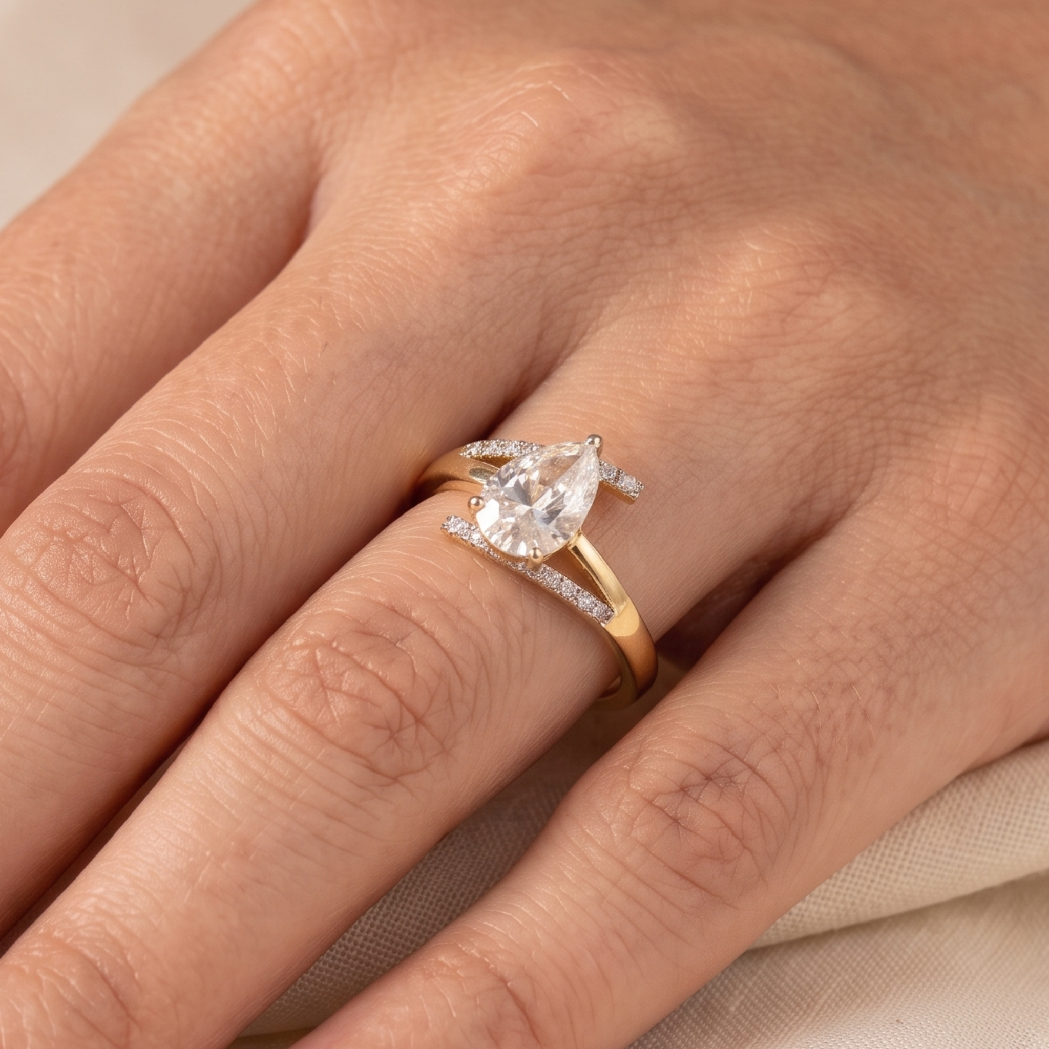 A close-up of a hand wearing a yellow gold ring with a pear-cut clear center stone and small side stones on a soft beige fabric background.