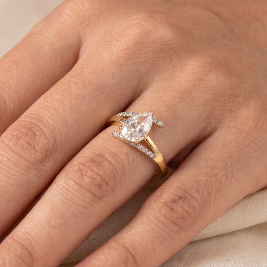 A close-up of a hand wearing a yellow gold ring with a pear-cut clear center stone and small side stones on a soft beige fabric background.