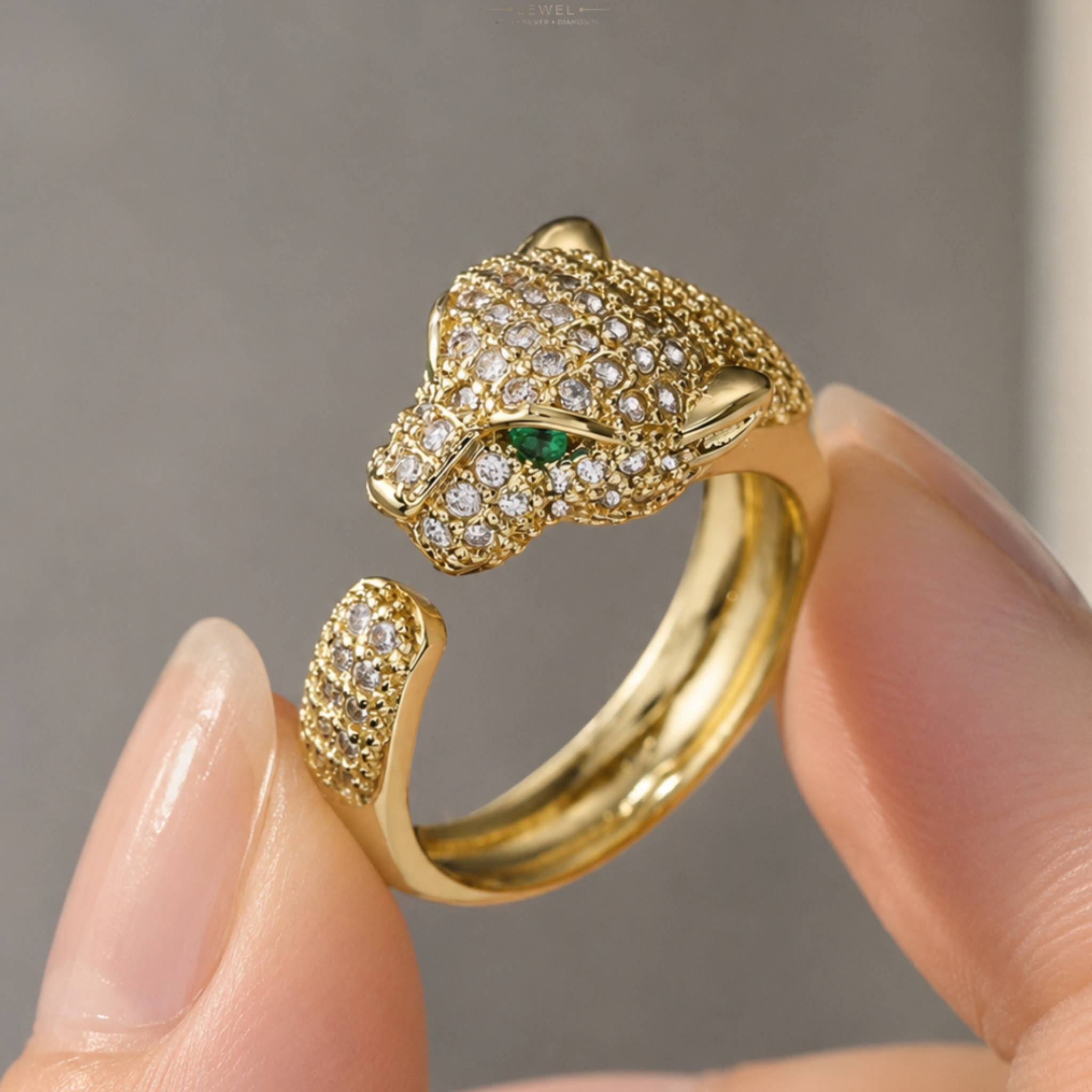 A gold-toned open ring designed like a panther or leopard head, covered in small clear stones with a small green eye accent, being held between fingers.