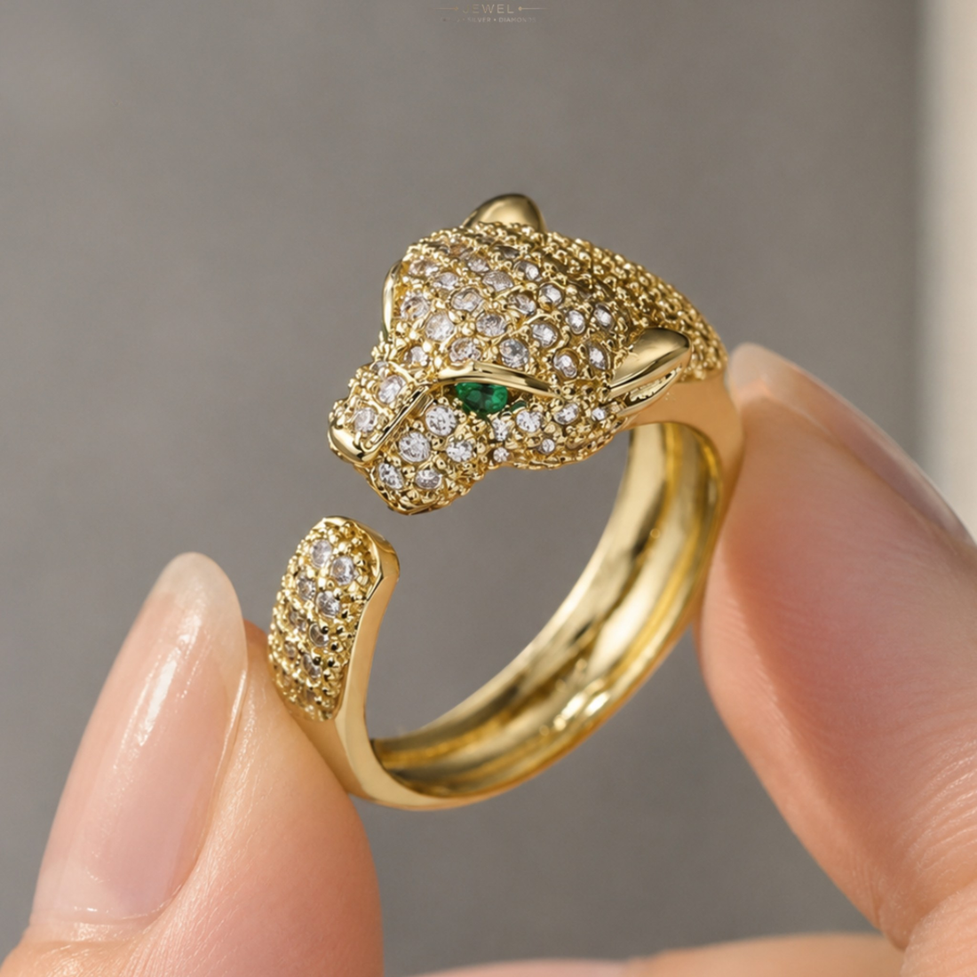 A gold-toned open ring designed like a panther or leopard head, covered in small clear stones with a small green eye accent, being held between fingers.