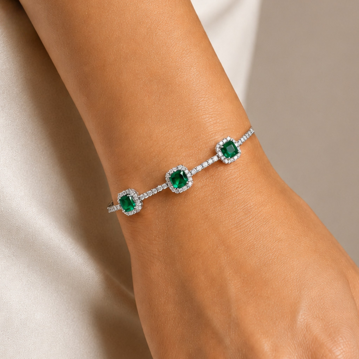 A close-up of a delicate silver-toned bracelet with three square-cut green emeralds, each surrounded by a halo of small clear stones, worn on a person's wrist.