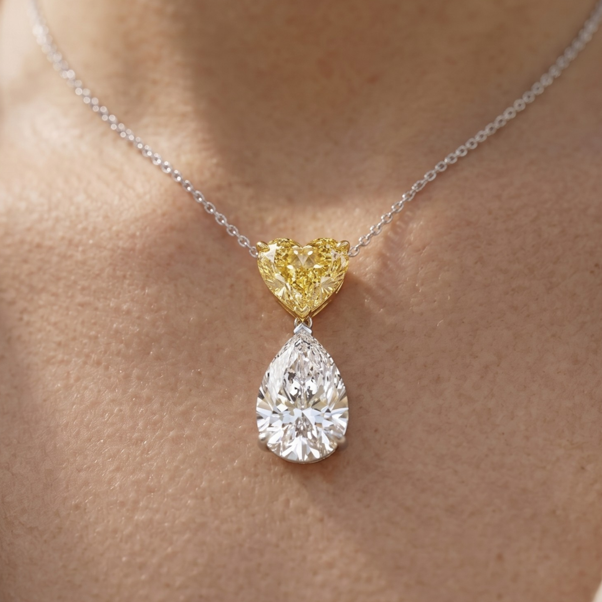 A close-up of a delicate necklace worn on skin, featuring a silver chain with a dual-stone pendant. The pendant has a small, heart-shaped yellow diamond at the top and a larger pear-cut clear diamond below in a silver setting.