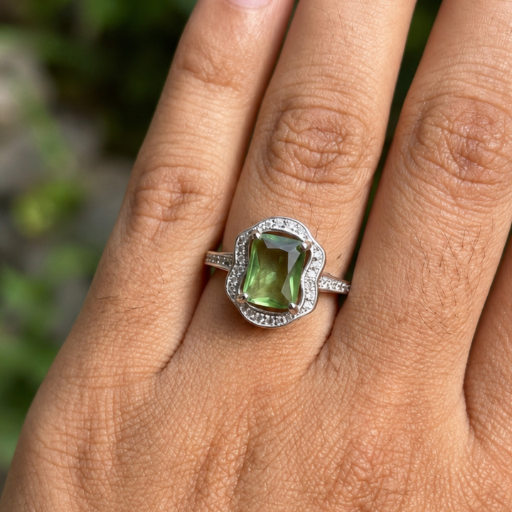 A silver ring with a rectangular green gemstone and a halo of small clear stones, shown worn on a person's finger against a blurred green background. The brand name 'GLAZE' is visible as a watermark in the top left corner.