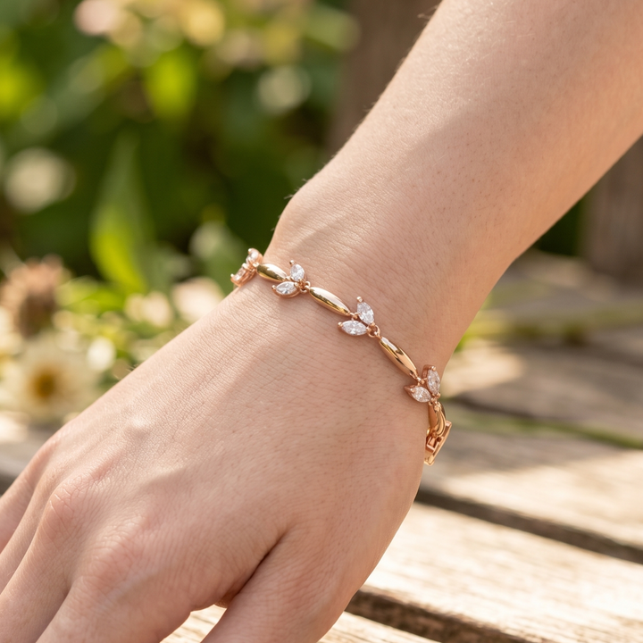 A delicate rose gold bracelet with small leaf-shaped crystal accents worn on a person's wrist, photographed outdoors against a blurred green garden and wooden surface.
