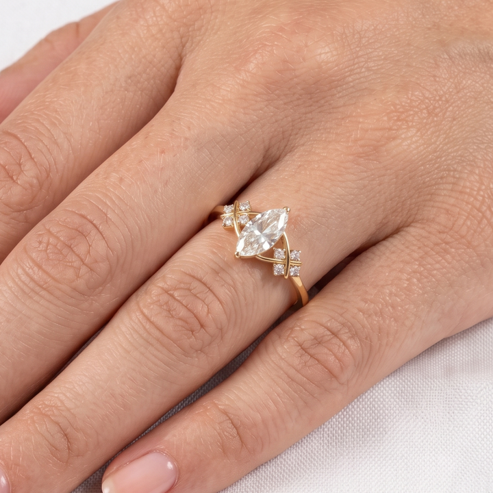 A gold ring with a marquise-cut clear center stone and small accent stones, shown worn on a person's finger against a light fabric background.