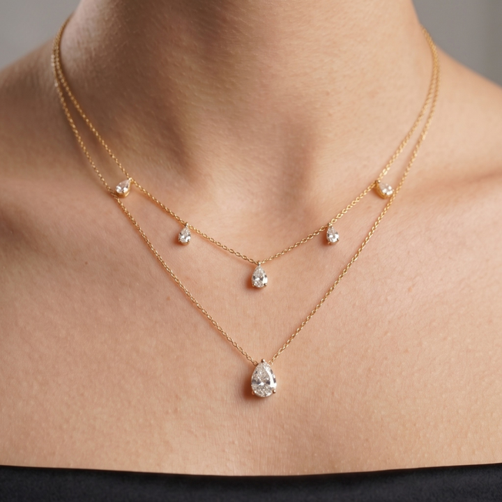 A close-up of a model's neck and upper chest wearing a delicate double-layered gold necklace with clear teardrop and round gemstones.