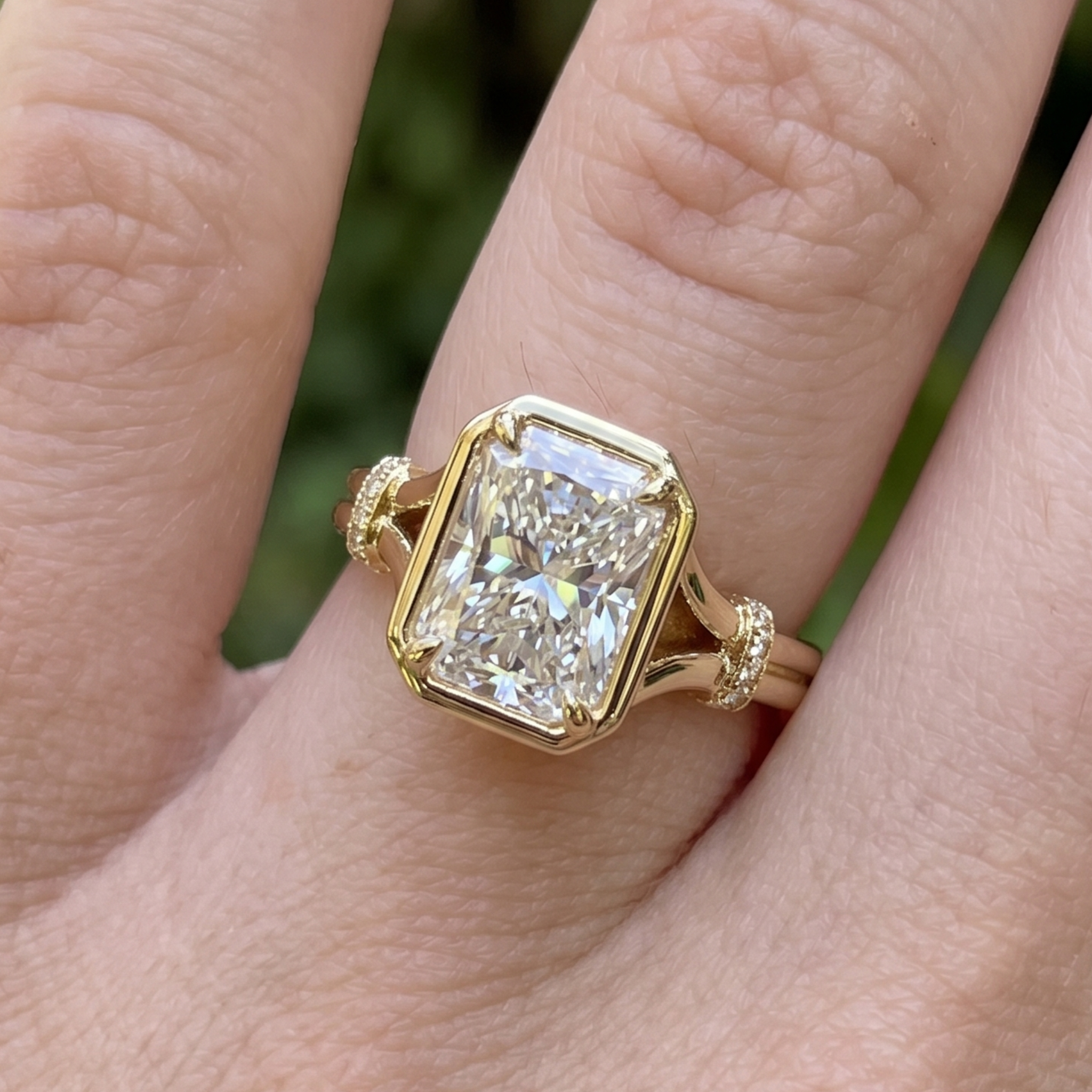 A close-up of a gold ring with a large rectangular clear gemstone, worn on a finger against a blurred green outdoor background.
