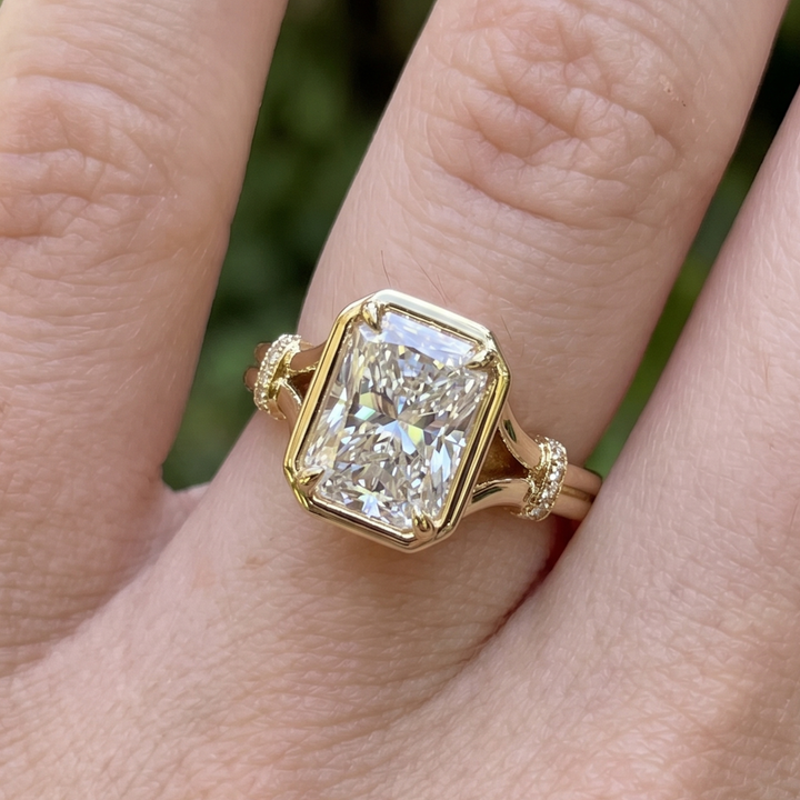 A close-up of a gold ring with a large rectangular clear gemstone, worn on a finger against a blurred green outdoor background.