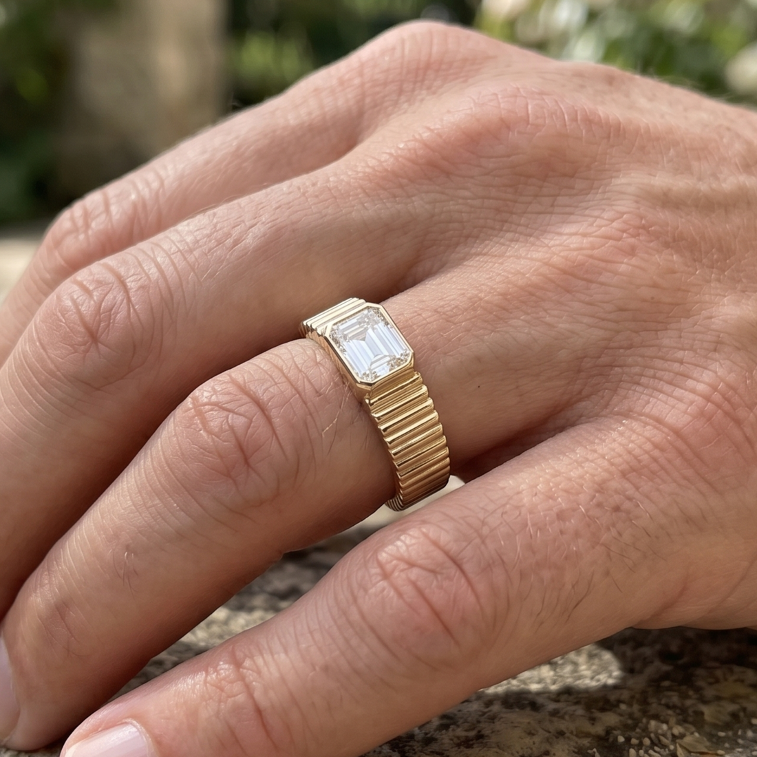 A gold ring with a rectangular baguette-cut clear gemstone, worn on a person's finger in an outdoor setting.