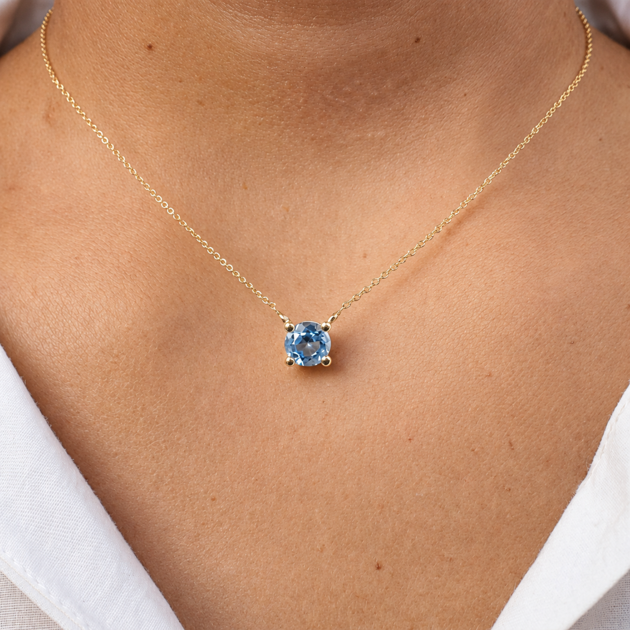 A delicate gold chain necklace with a round-cut light blue gemstone pendant, shown worn on a person's neck over a white collared shirt.