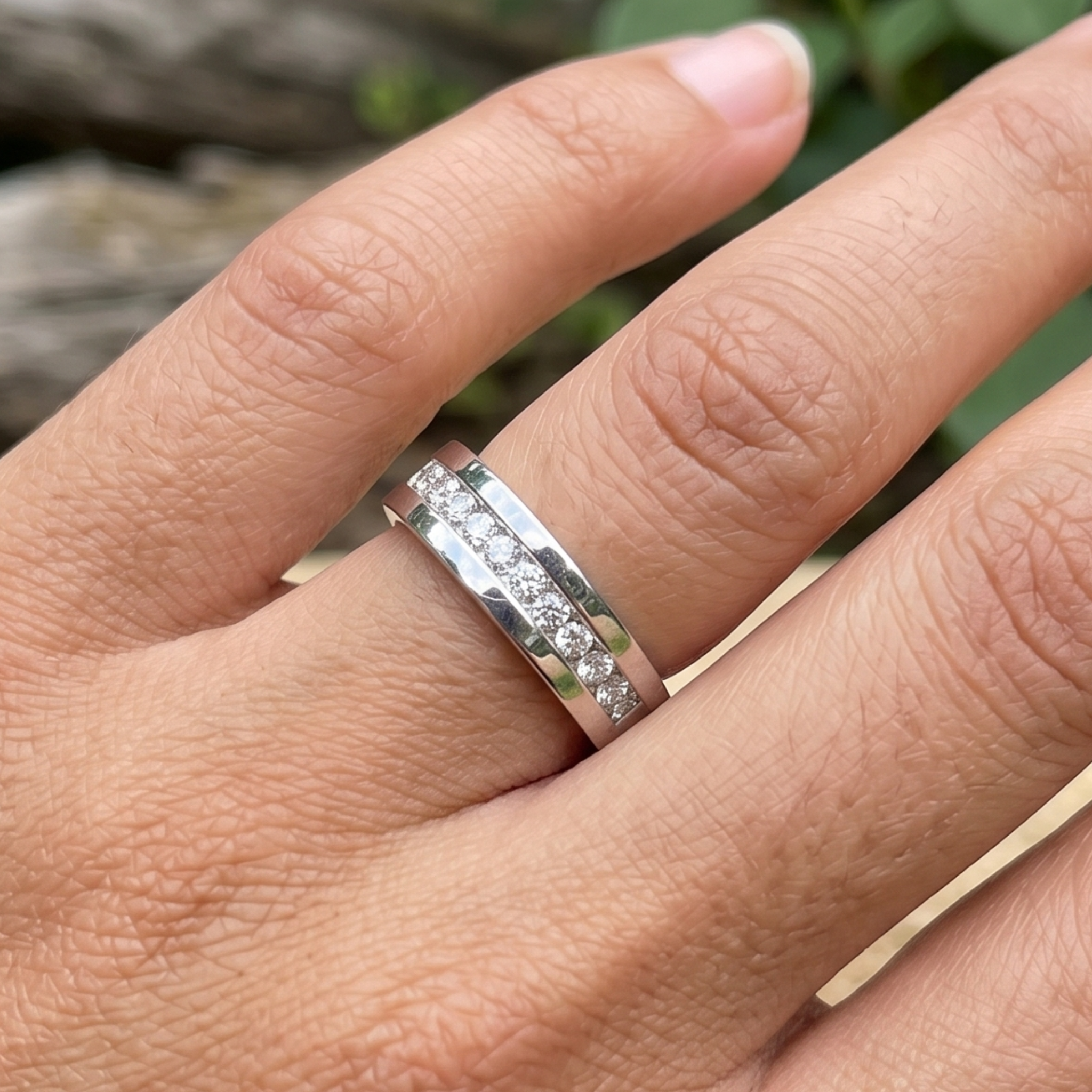A silver band ring with a channel-set row of small clear stones, shown worn on a person's finger against a blurred outdoor background.