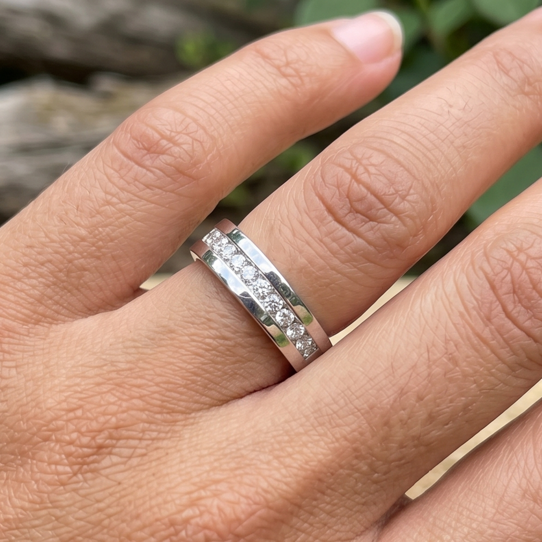 A silver band ring with a channel-set row of small clear stones, shown worn on a person's finger against a blurred outdoor background.