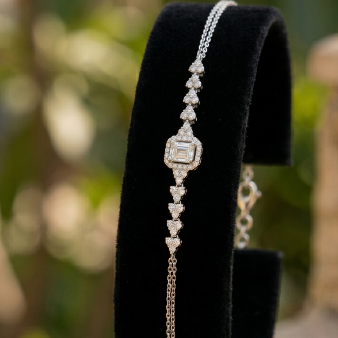 A silver-toned bracelet with a central square diamond and heart-shaped diamond accents, displayed on a black velvet jewelry stand against a blurred green background.