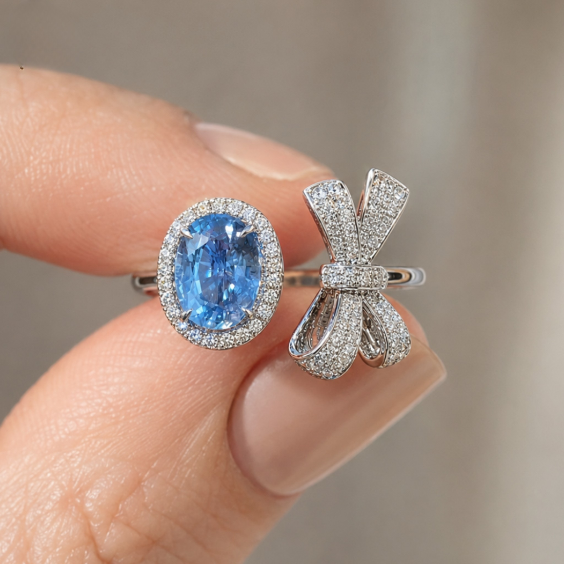 A close-up of a hand holding two rings: one featuring an oval-cut blue sapphire gemstone surrounded by a halo of small clear stones, and the other a silver-toned bow-shaped ring paved with clear stones.