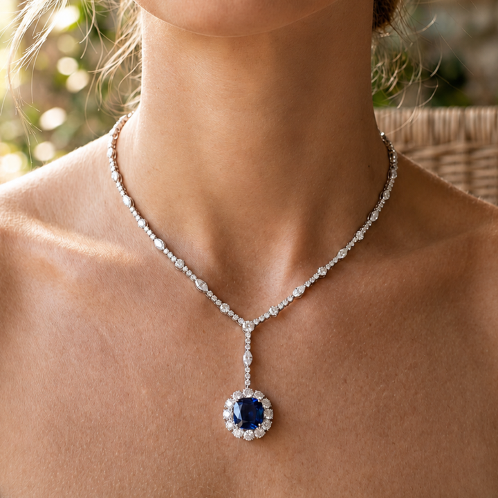 A close-up of a model wearing a silver-toned Y-shaped necklace with a round deep blue gemstone pendant surrounded by clear stones.