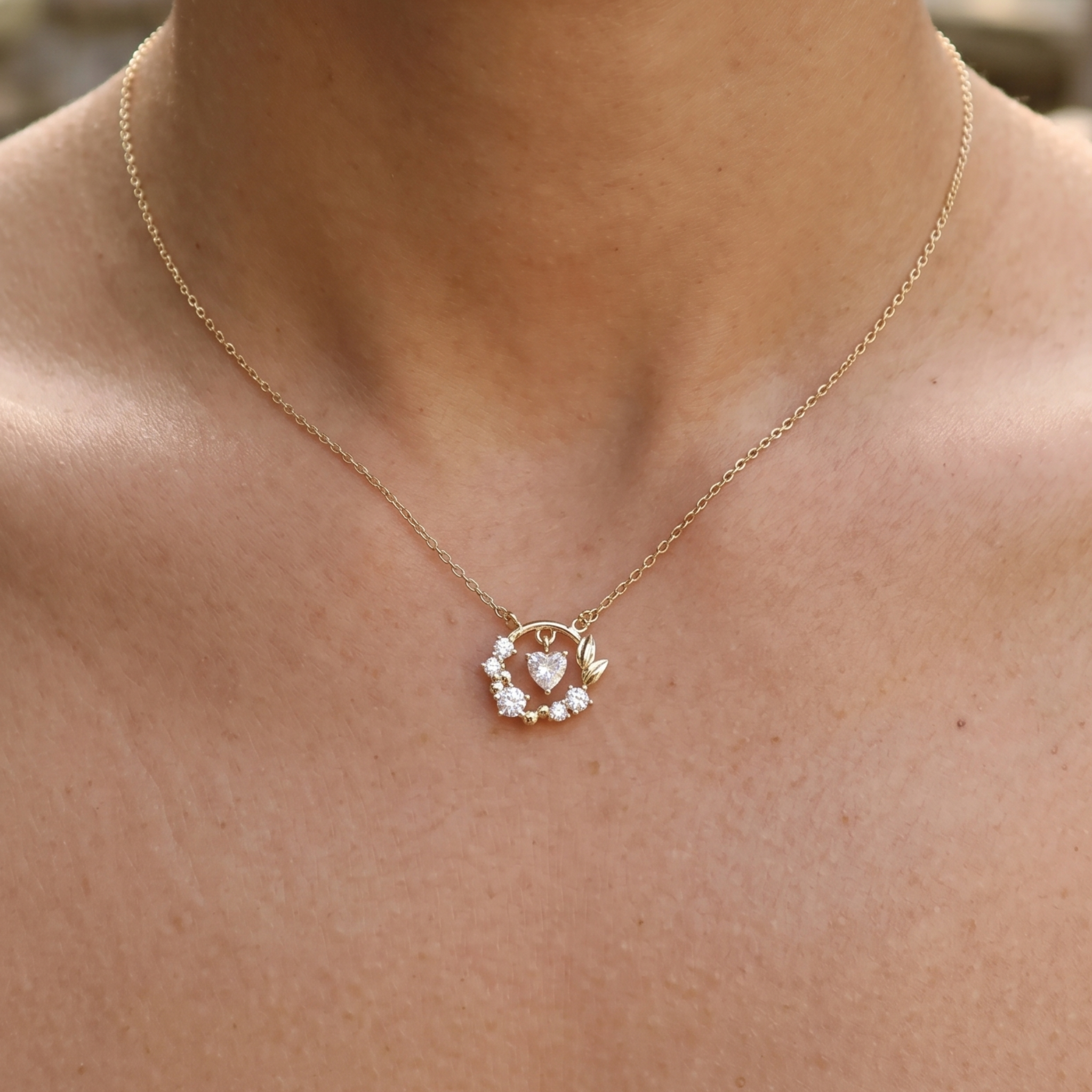 A delicate gold chain necklace with a small circular pendant featuring a central heart and clear stones, shown worn on a person's neck and upper chest.