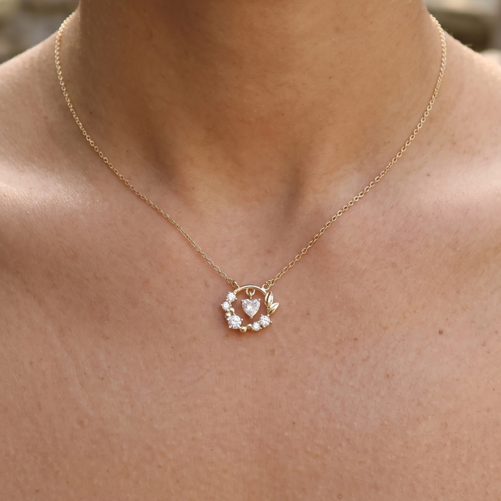 A delicate gold chain necklace with a small circular pendant featuring a central heart and clear stones, shown worn on a person's neck and upper chest.
