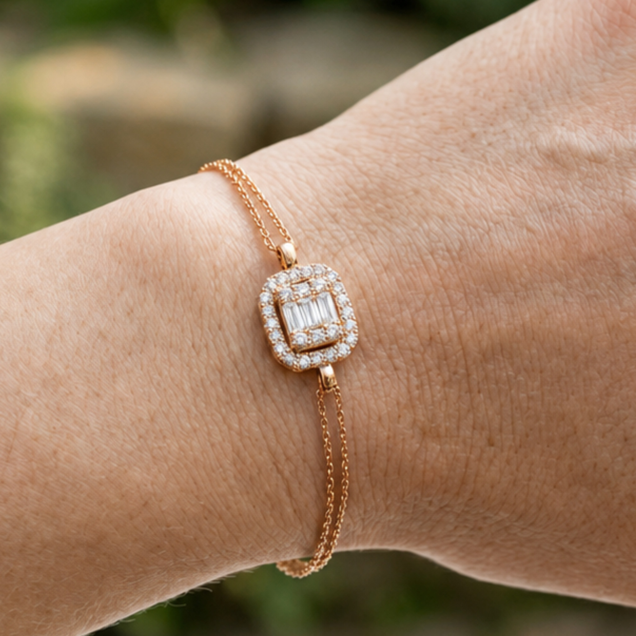A close-up of a delicate rose gold bracelet with a rectangular baguette diamond center and a surrounding diamond halo, worn on a person's wrist.
