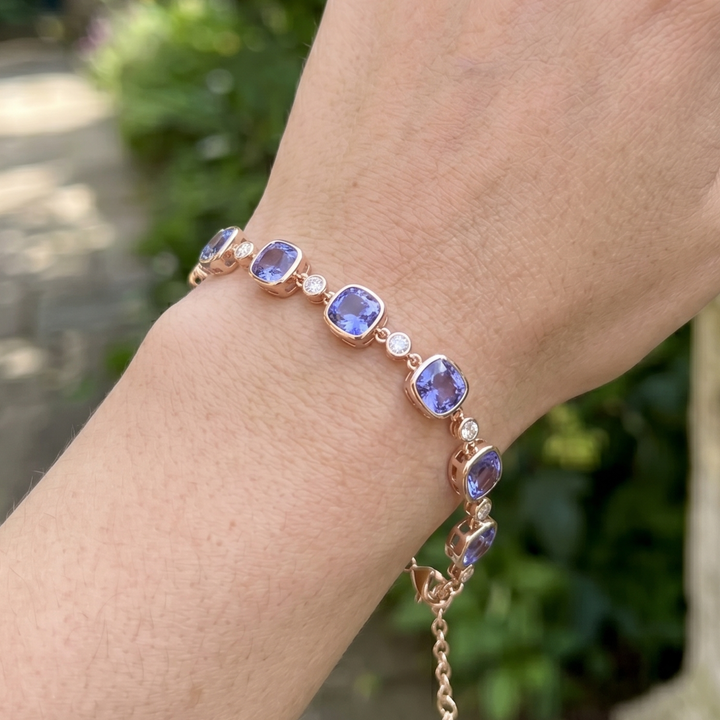 A close-up of a rose gold bracelet with lavender tanzanite gemstones and small clear accent stones, worn on a person's wrist against a blurred outdoor background.
