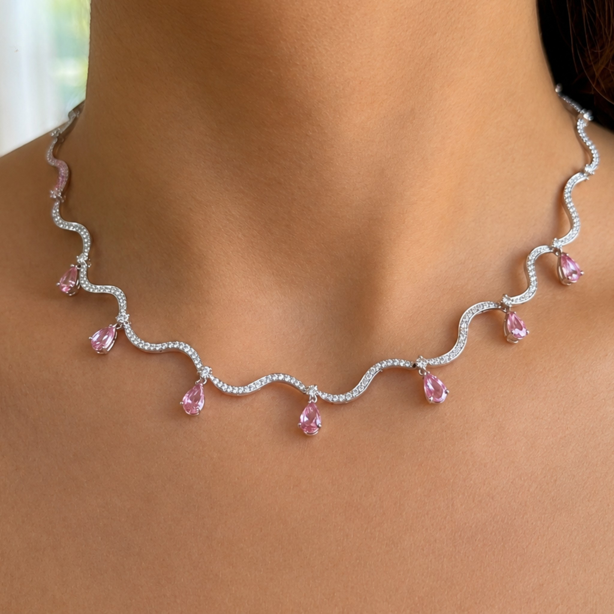 A close-up of a woman's neck and upper chest wearing a wavy silver-toned necklace with small clear stones and pink teardrop gemstones.
