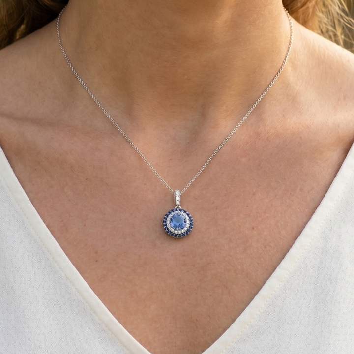 A close-up of a woman's neck and chest wearing a delicate silver chain necklace with a round pendant featuring a light blue center surrounded by a dark blue halo and small clear stones.