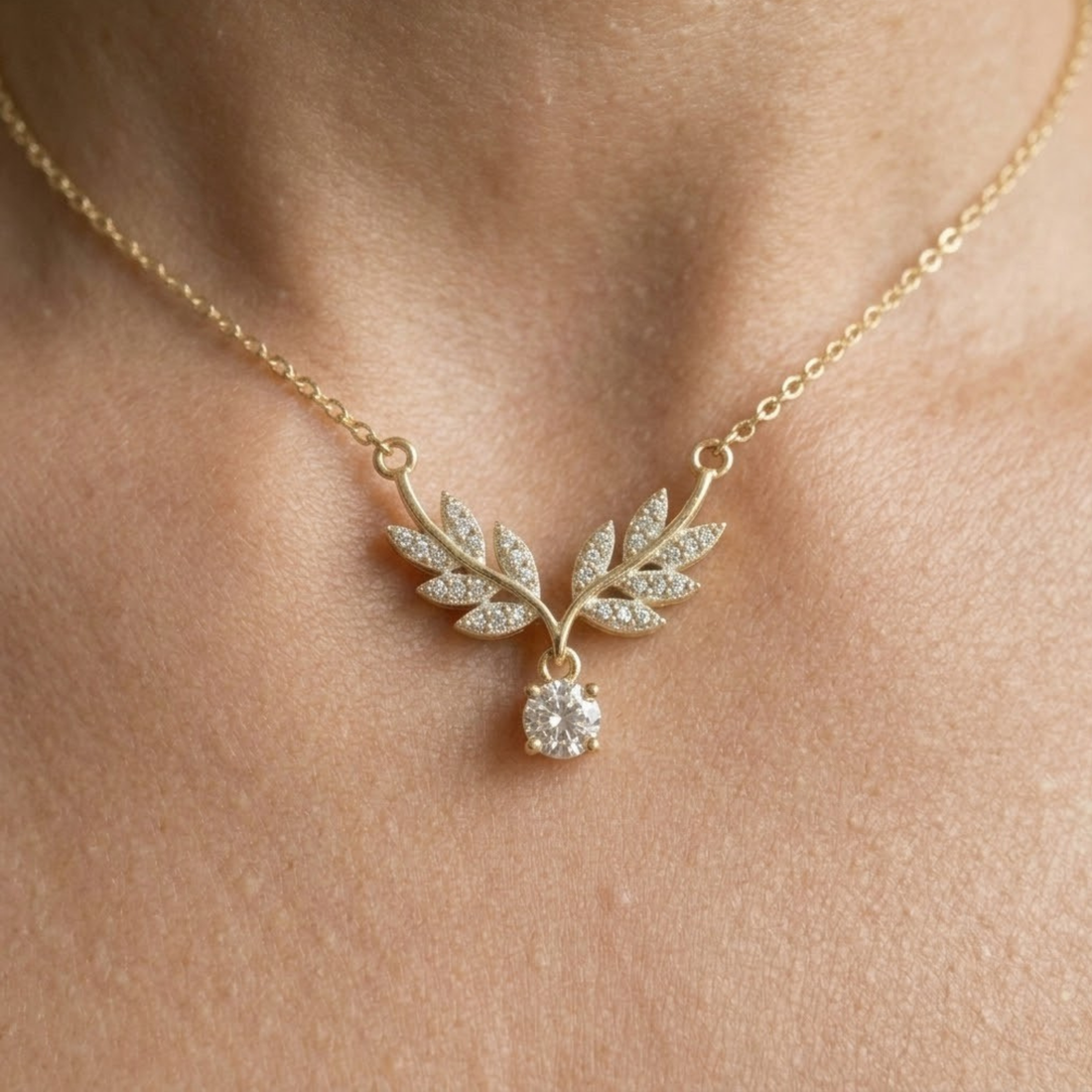 A delicate gold necklace with a leaf-shaped pendant paved with small clear stones and a round clear gemstone drop, shown worn on a person's neck.