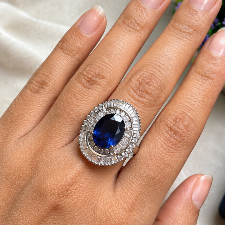 A close-up of a hand wearing a large oval ring with a deep blue center stone and a silver-toned halo setting with small clear stones.