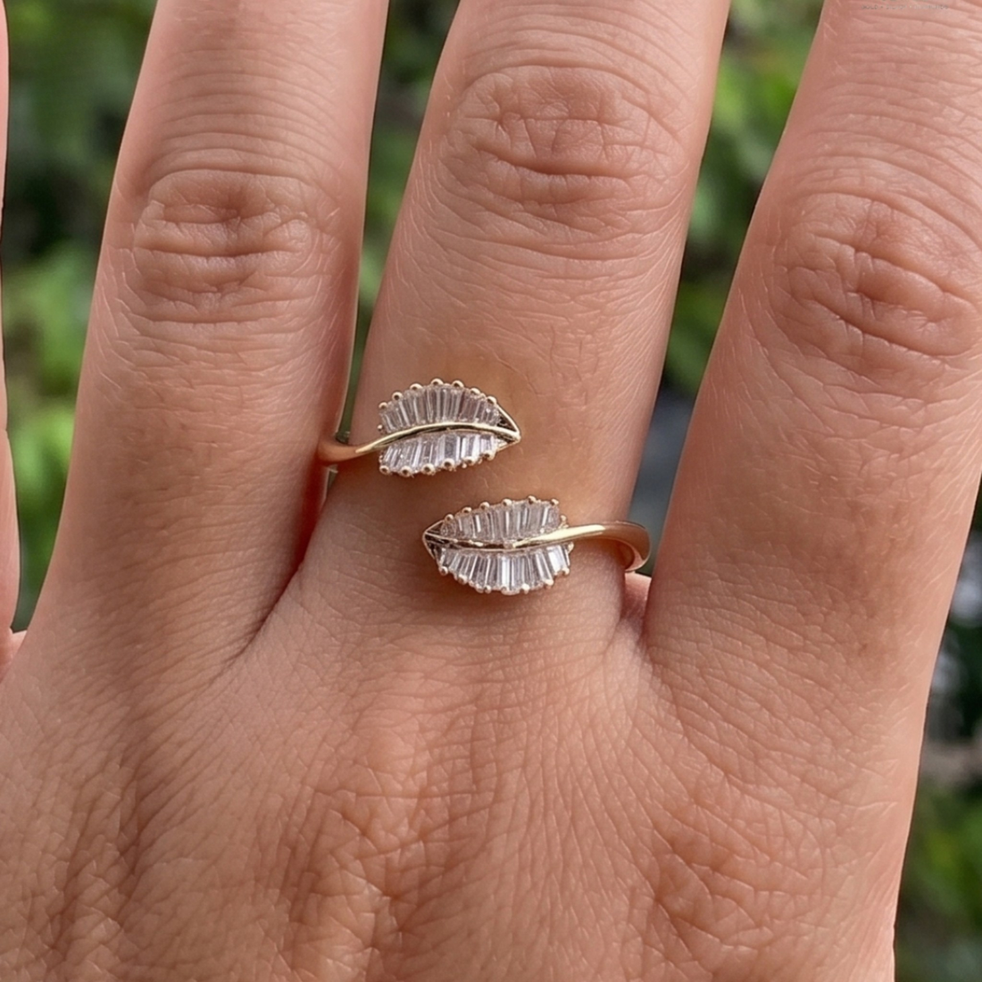 A close-up of a hand wearing two small leaf-shaped rings with clear faceted stones on a gold-tone band, against a blurred green outdoor background.