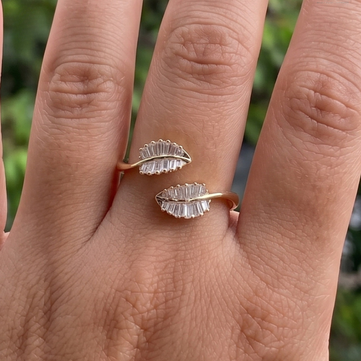 A close-up of a hand wearing two small leaf-shaped rings with clear faceted stones on a gold-tone band, against a blurred green outdoor background.