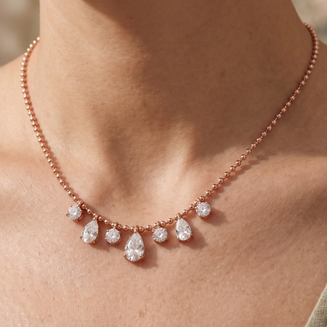 A close-up of a person's neck and collarbone wearing a delicate rose gold necklace with five dangling pear-cut clear gemstones.
