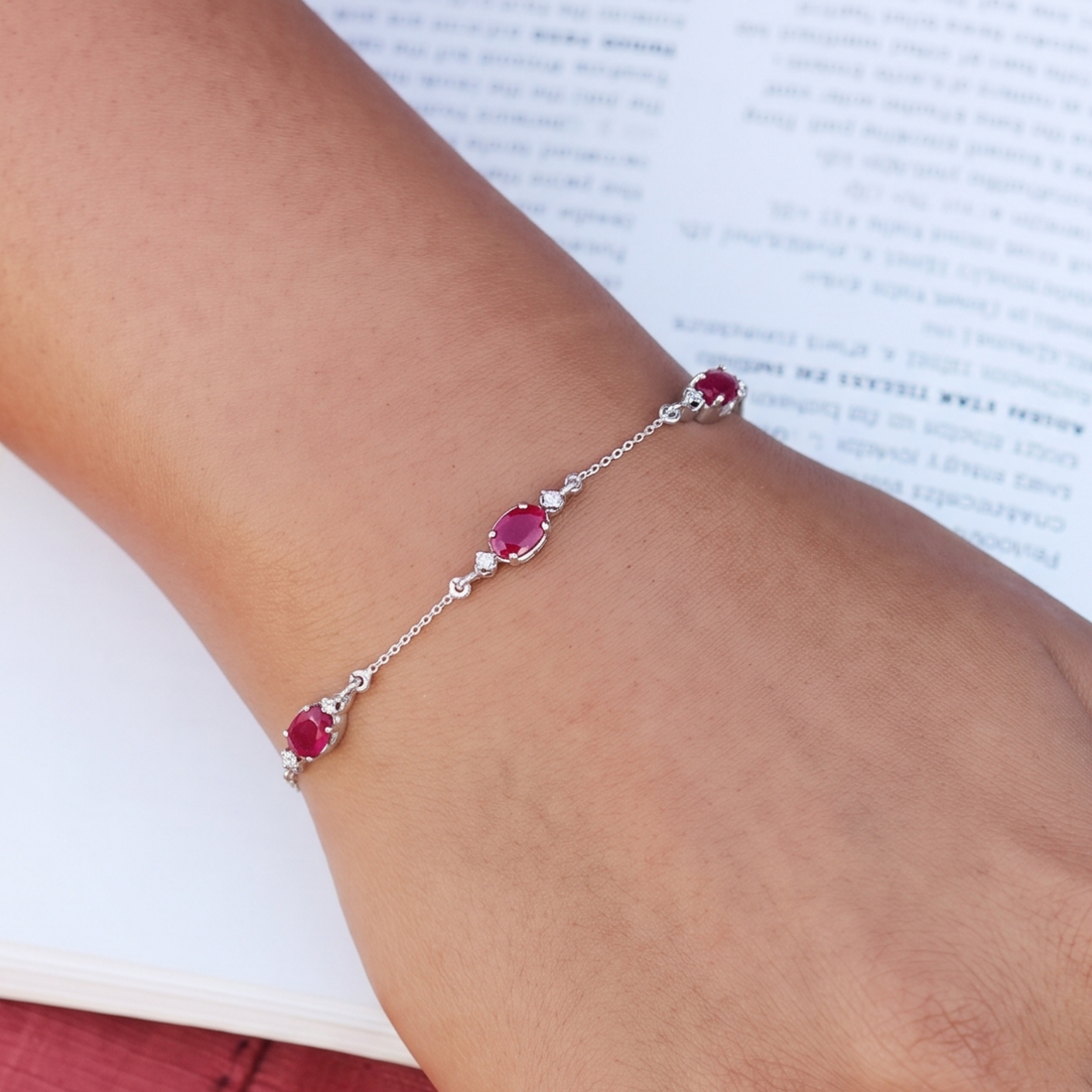 A delicate silver chain bracelet with three oval pinkish-red gemstones and small clear accent stones, shown worn on a person's wrist over an open book.