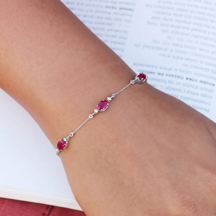 A delicate silver chain bracelet with three oval pinkish-red gemstones and small clear accent stones, shown worn on a person's wrist over an open book.