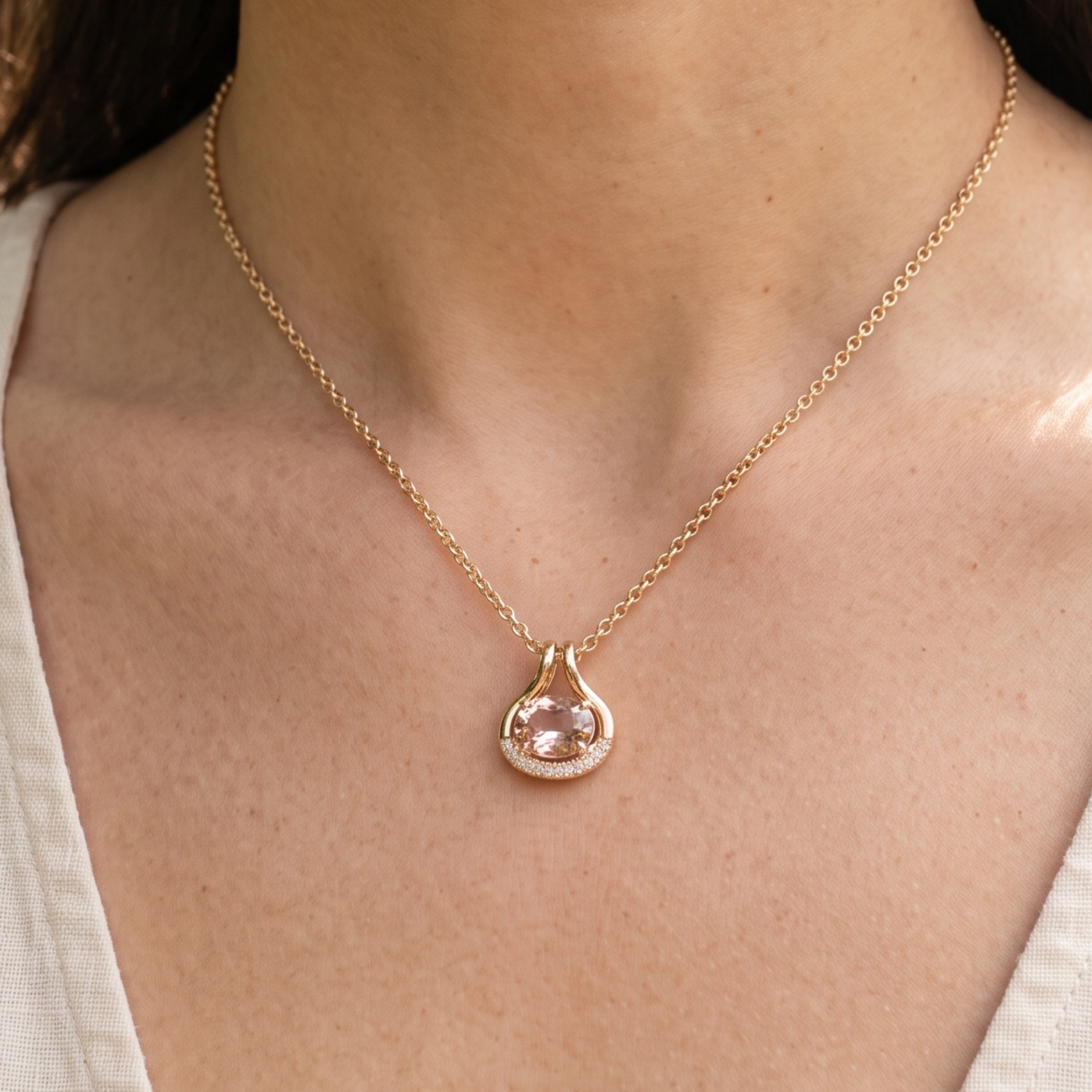 A close-up of a model wearing a delicate gold chain necklace with a teardrop-shaped pendant featuring a light pink gemstone surrounded by small clear stones.