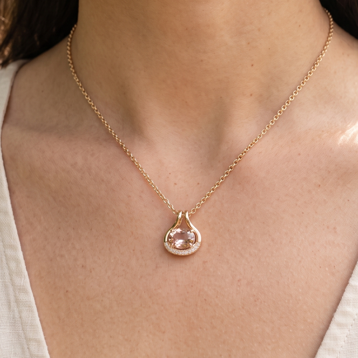 A close-up of a model wearing a delicate gold chain necklace with a teardrop-shaped pendant featuring a light pink gemstone surrounded by small clear stones.