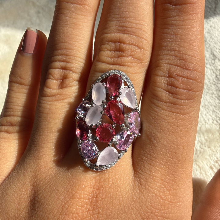 A close-up of a hand wearing a large oval cluster ring with pink and light lavender teardrop-shaped gemstones and a silver-toned setting.
