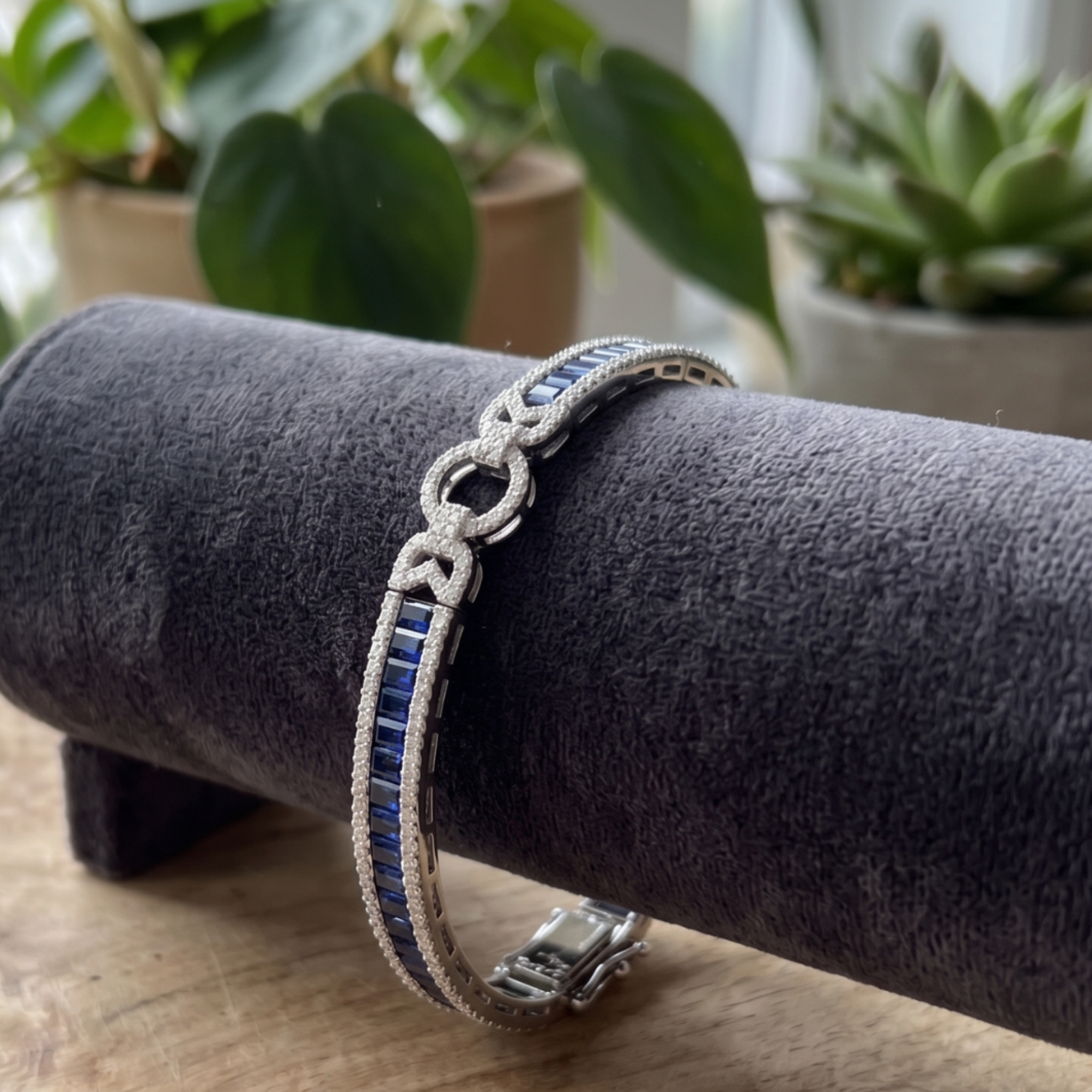 A silver-toned bracelet with a row of rectangular blue stones, displayed on a dark gray velvet jewelry stand over a wooden surface, with blurred green plants in the background.