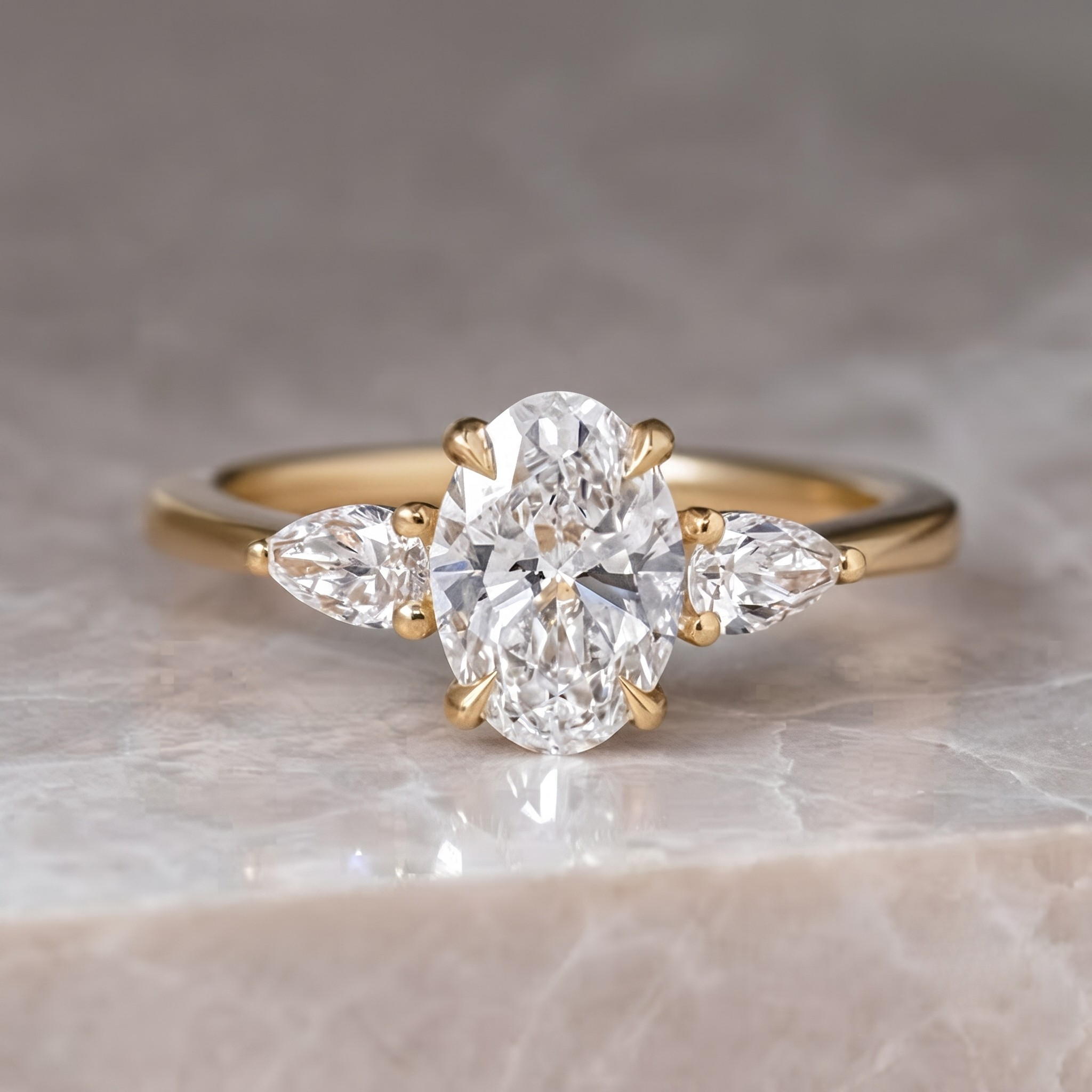 A yellow gold engagement ring featuring a large oval center diamond flanked by two smaller pear-cut diamonds, displayed on a light marble surface.