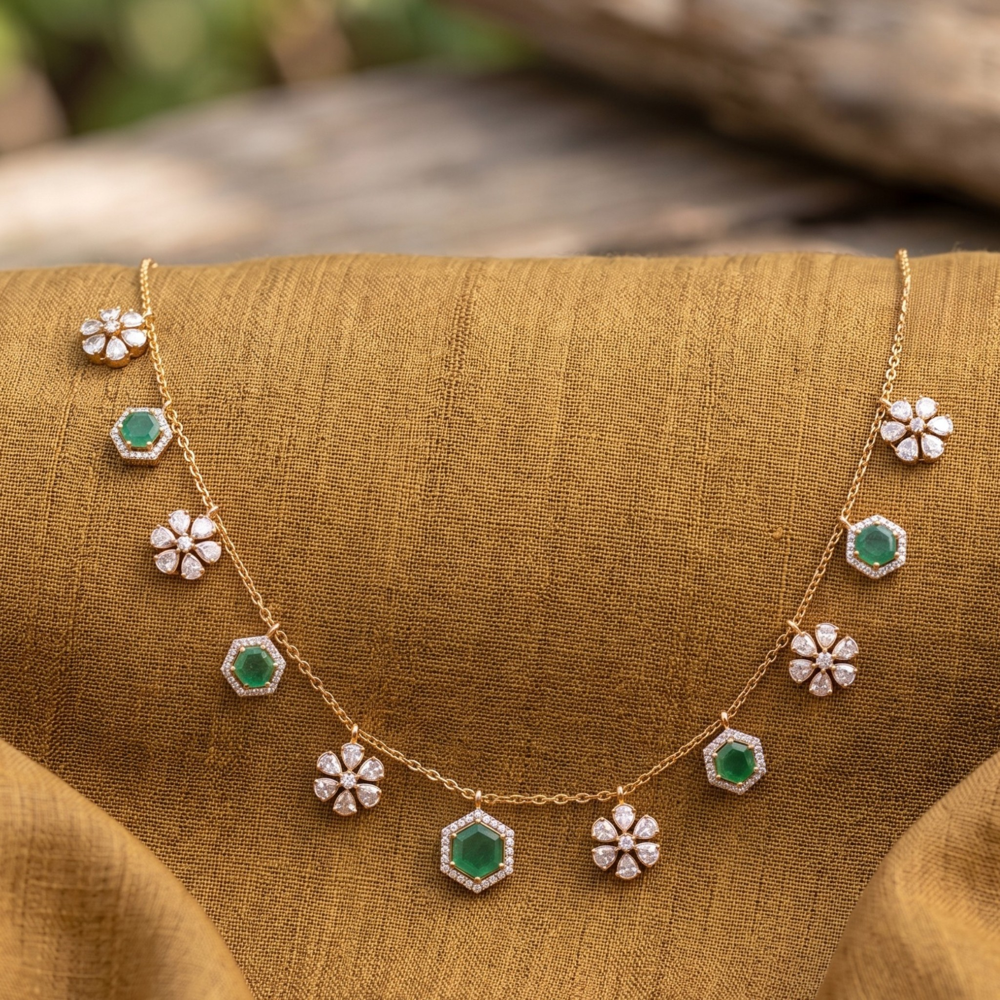 A gold necklace with alternating green hexagonal and white floral gemstone motifs, displayed on a textured mustard-brown fabric surface.