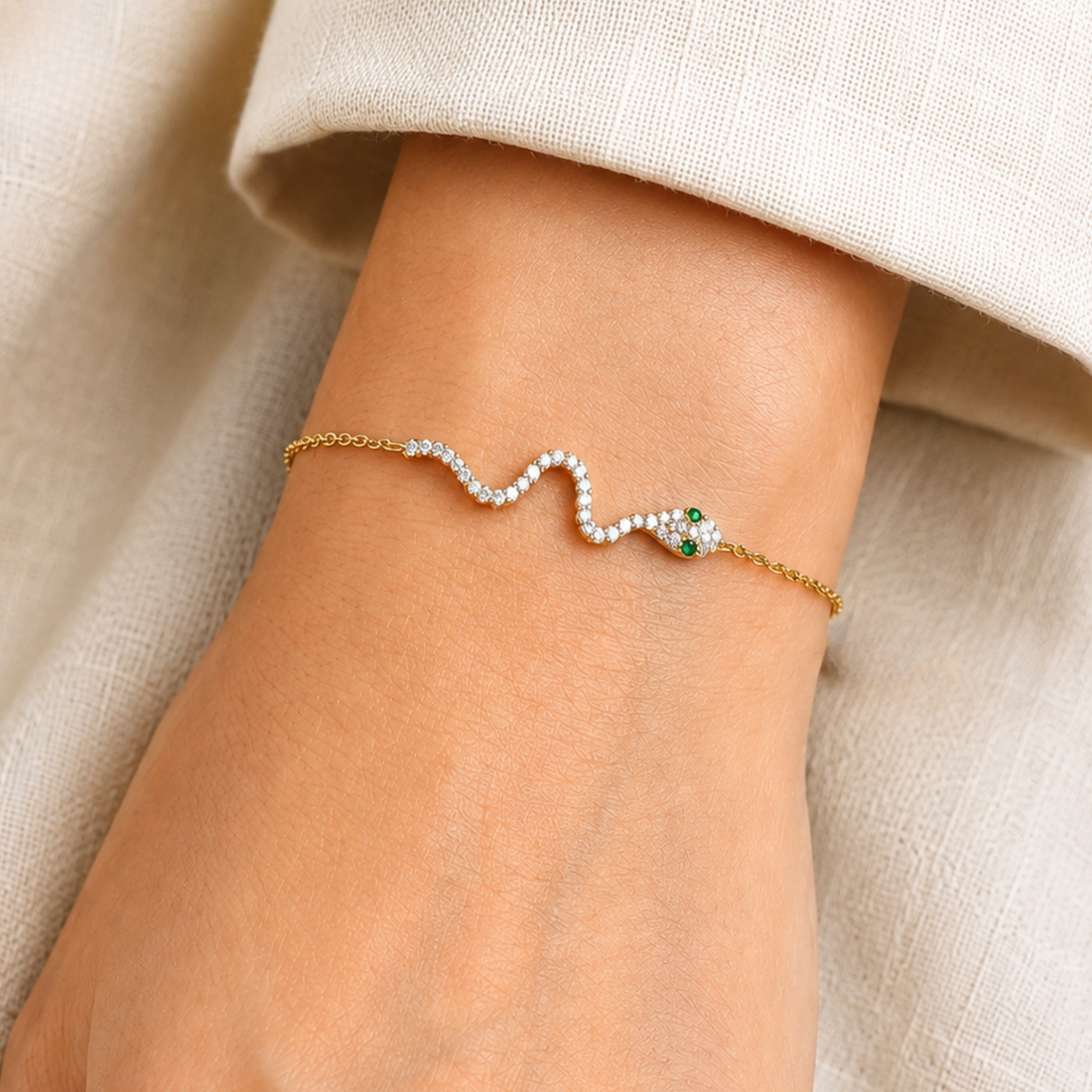 A delicate gold chain bracelet with a wavy design, featuring a row of small white stones and a green-and-white snake head motif on the wrist, shown on a model's hand with a beige sleeve.
