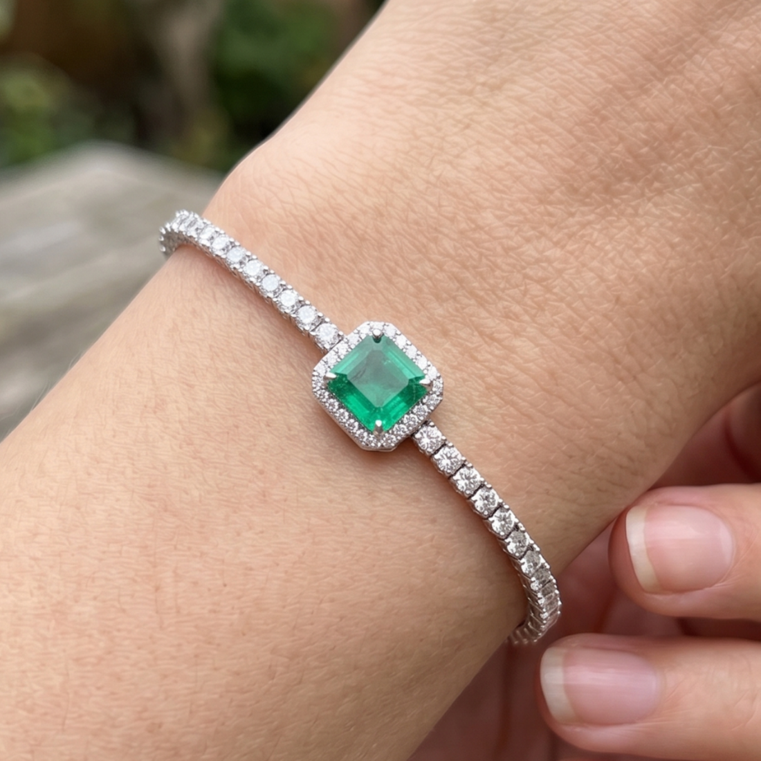 A close-up of a silver-toned band bracelet worn on a wrist, featuring a square-cut green emerald center stone surrounded by a halo of small clear stones, with additional clear stones lining the band.