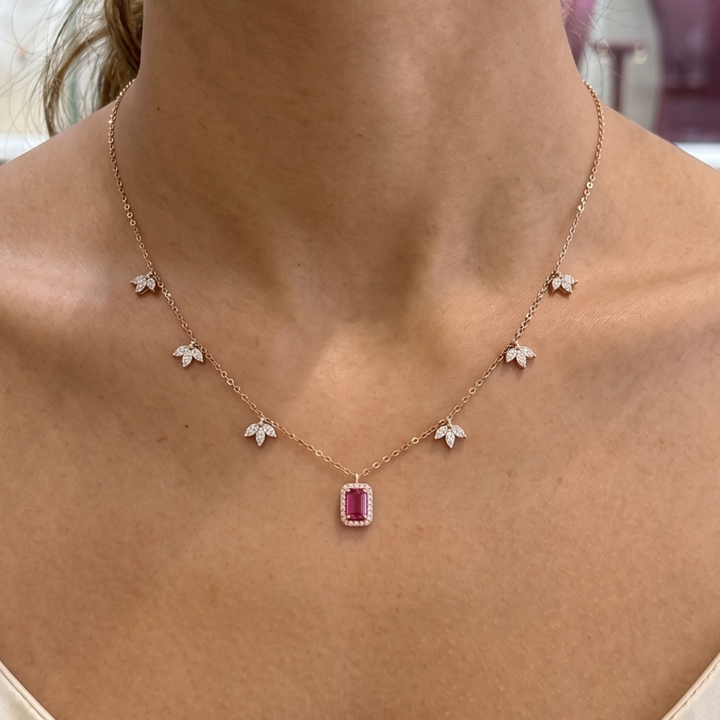 A close-up of a model's neck and upper chest wearing a delicate rose gold necklace. The necklace features a central rectangular pink gemstone pendant surrounded by small white flower-shaped charms and a fine chain.