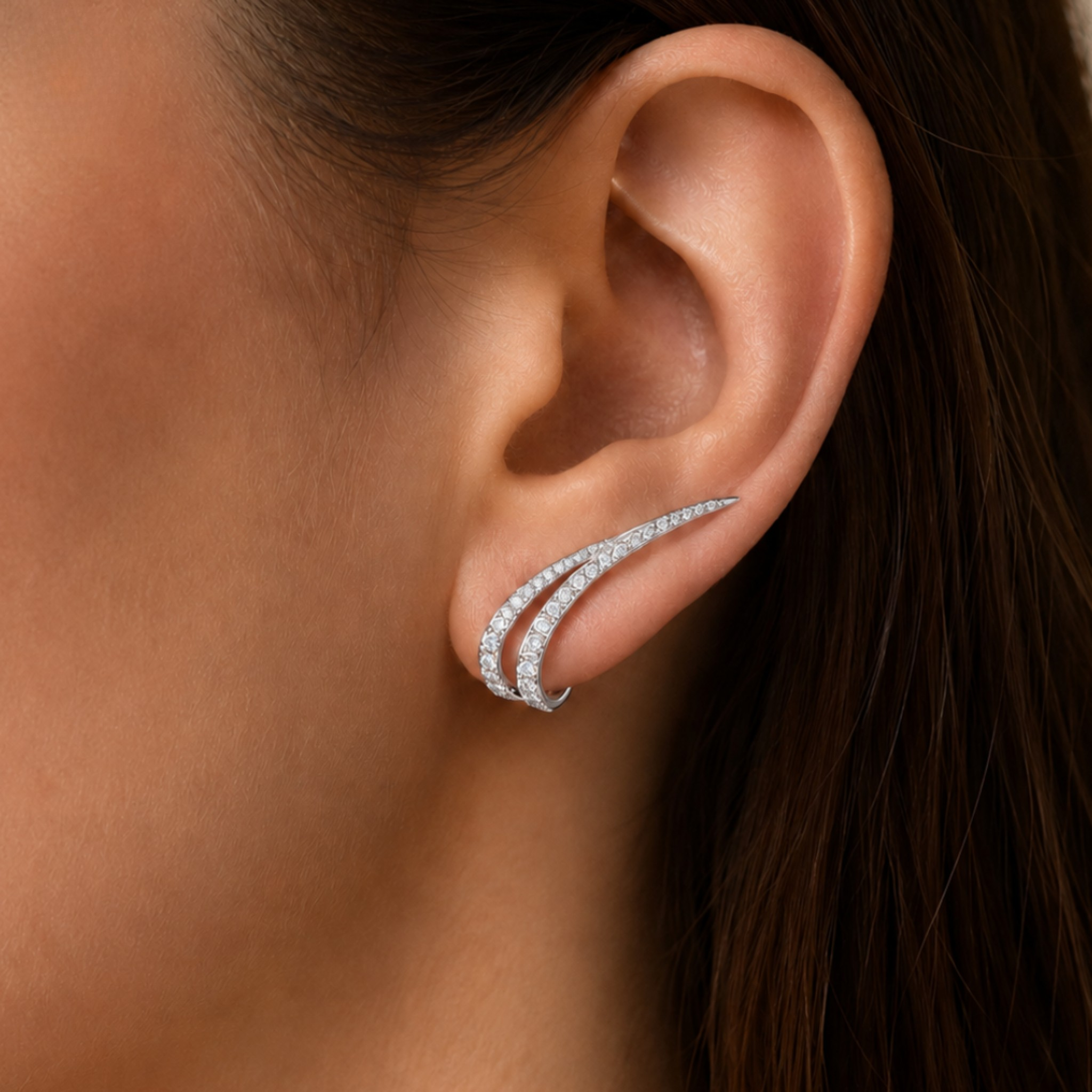 A close-up of a woman's ear wearing a curved, double-band ear cuff earring set with small clear stones in a silver-toned metal.